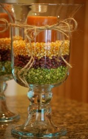 a glass candleholder filled with peas, beans and corn and with a bright pillar candle on top is a bright and rustic fall decoration