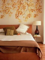 a fall-colored bedroom with a leafy wall, an orange bed, muted color pillows, a table lamp with a brown glass base
