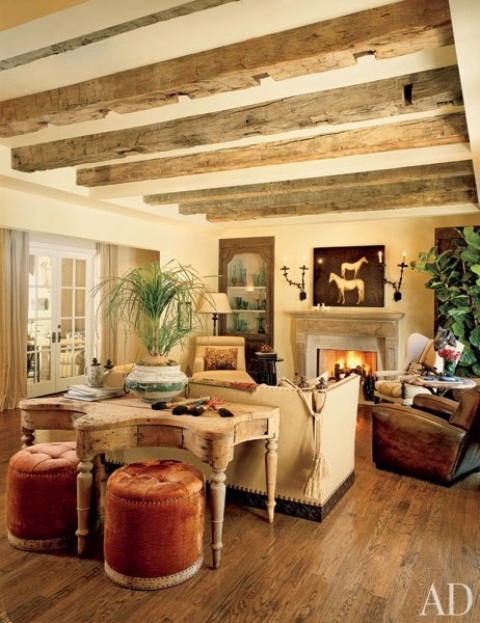 a refined vintage barn living room with wooden beams, a beautiful fireplace, neutral and brown seating furniture, a vintage table, rust-colored stools and potted greenery