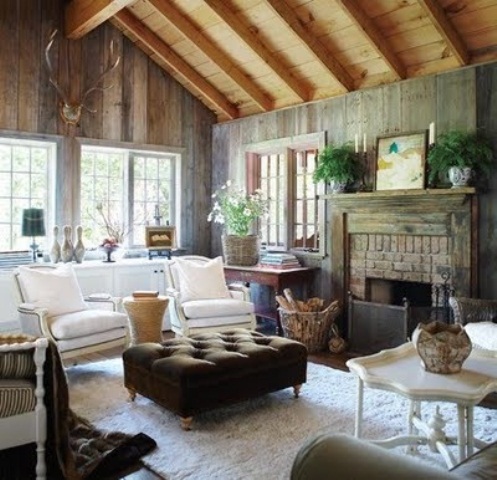 a barn living room with reclaimed wood walls, a fireplace clad with brick, vintage white seating furniture, a low tufted ottoman, potted greenery and blooms is amazing