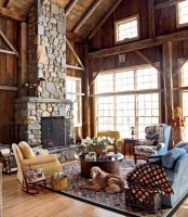 a barn living room clad with wood and with a wooden ceiling, a fireplace clad with stone, blue and yellow seating furniture, printed textiles and potted blooms