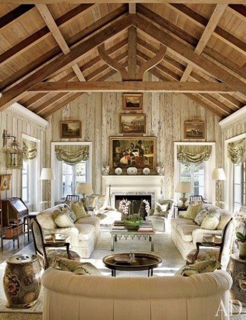 a vintage barn living room with wooden beams and a ceiling,w ith whitewashed walls, neutral vintage furniture, dark stained tables and a desk, baskets and curtains