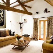 a welcoming barn living room with wooden beams, tan-colored seating furniture, bright and printed textiles, a stained wooden table and a coat of arms
