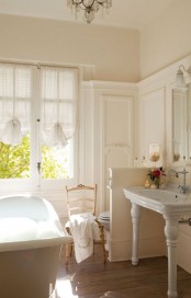 a light-filled farmhouse bathroom done in creamy shades, a clawfoot bathtub, elegant curtains-shades, a vintage vanity