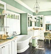a green farmhouse bathroom with an oval tub, creamy vanities and a storage unit, a vintage chair and mirrors