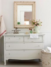 a vintage farmhouse bathroom with a white sideboard, a shabby chic frame mirror, a fabric basket for storage