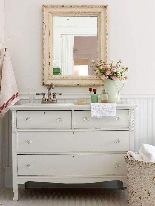 a vintage farmhouse bathroom with a white sideboard, a shabby chic frame mirror, a fabric basket for storage a vintage farmhouse bathroom with a white sideboard, a shabby chic frame mirror, a fabric basket for storage