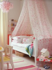 a lovely girl’s room with neutral walls and a floor, a white bed with floral bedding, a pink floral canopy and a wall-mounted shelf, lovely toys and chairs