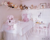 a neutral and light pink girl’s room with a small metal bed with neutral bedding, a play dining set, an open long shelf with all the toys stored and displayed