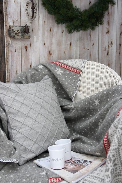 a white wicker chair with an embroidered grey blanket, a matching textured pillow, some mugs and an evergreen wreath on the wall