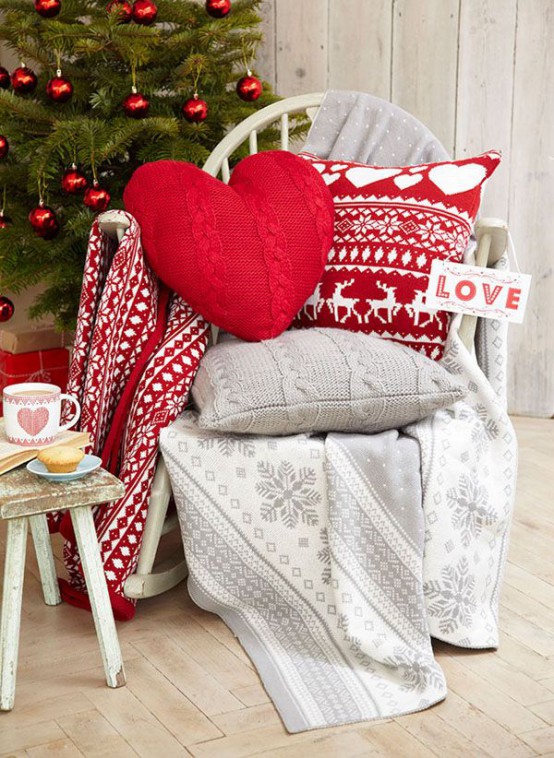 a chair styled for Christmas with knit pillows, grey and red printed blankets and pillows, a Christmas tree with red ornaments in the backdrop