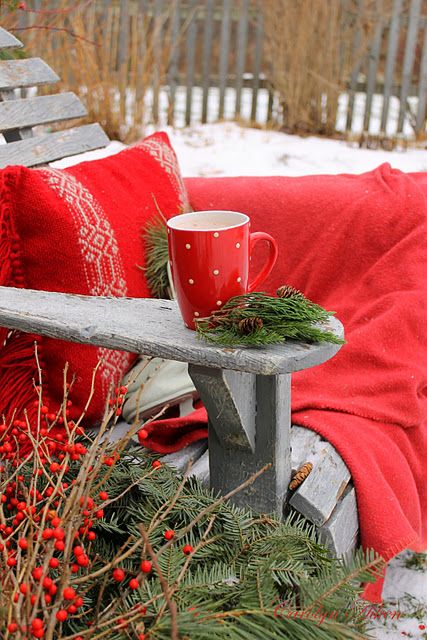 a simple wooden grey chair styled with a bold red blanket and a pillow, some evergreens with berries and a red mug is a great piece to enjoy winter outdoors