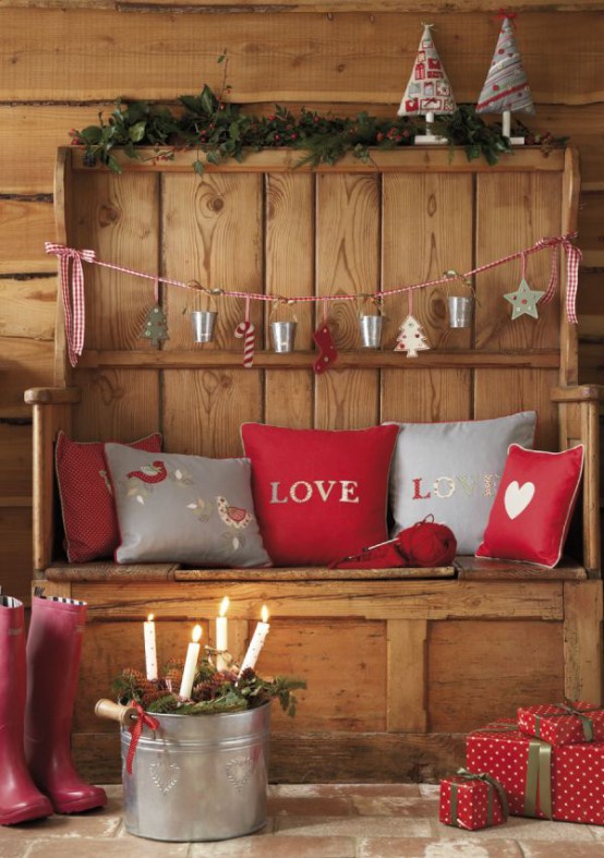 a rustic wooden bench decorated with grey and red printed Christmas pillows, with a grey and red garland, red gift boxes and a bucket with candles