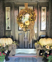 neutral oversized heirloom pumpkins in urns, neutral blooms, a wreath fo neutral pumpkins, blooms, herbs and greenery make up a cool rustic Thanksgiving front door