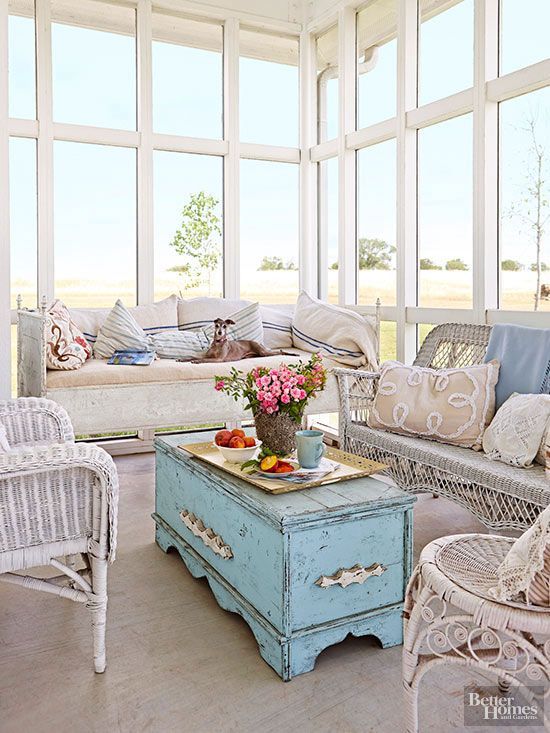 a coastal sunroom with a wooden sofa with striped pillows, white wicker furniture, a light blue chest as a coffee table and embroidered pillows