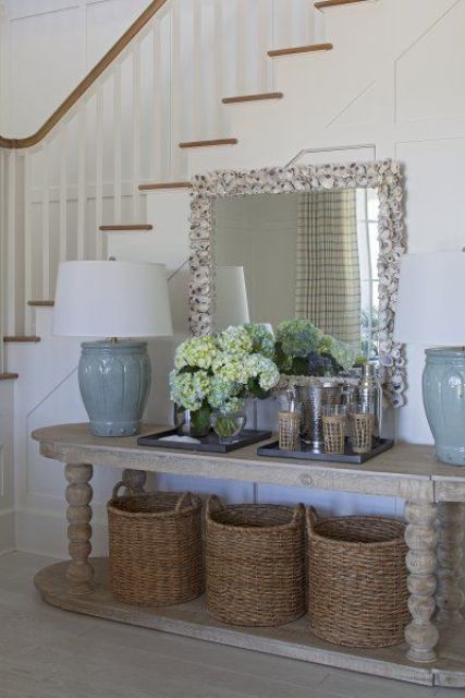 an elegant beach console table with wicker baskets, blue table lamps, blooms and a mirror in a seashell frame