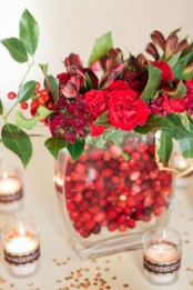 a glass vase filled with cranberries, with red and burgundy blooms and surrounded with candles is a beautiful Christmassy arrangement with plenty of color