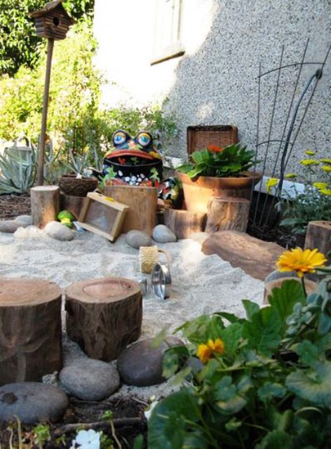 a fun and cute outdoor playground with plants, stumps, sand, pebbles, blooms and some garden figurines