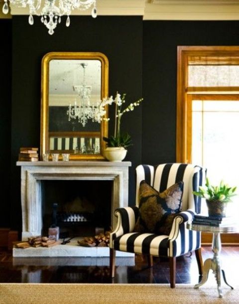 a sophisticated living room with black walls, a non-working fireplace and a large mirror, a black and white chair, a crystal chandelier