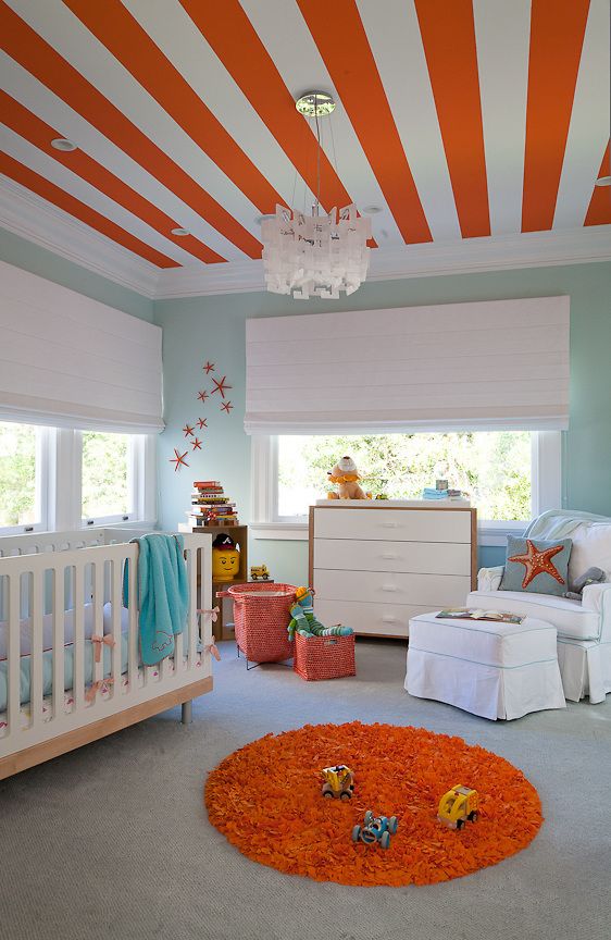 a beach nursery done in light blue, orange and white, with a bold striped ceiling, neutral furniture, an orange rug and some pretty toys