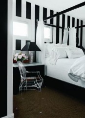 a sophisticated black and white bedroom with a stripe accent wall, a black canopy bed with white bedding, a black and white dresser and a black table lamp