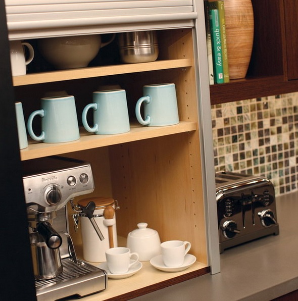 a storage cabinet with a coffee machine, mugs and cups and sugar is a mini coffee station with a shutter sliding down