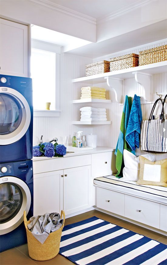 a coastal farmhouse laundry with white open shelves, baskets for storage, with a bench with drawers, a navy washing machine and a dryer, a striped rug, yellow and white towels