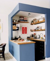 a small kitchen placed in a blue cube, with built-in black cabinets, light-colored woodne countertops and open shelving