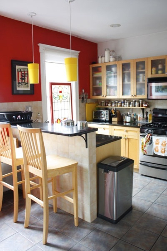 a small bright kitchen with a statement red wall, yellow pendant lamps, light stained furniture and a tall countertop for eating