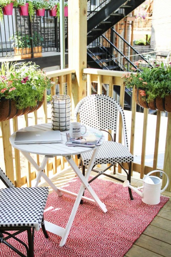 a small and bright summer balcony with dark wooden chairs, a white table, a bright rug and potted greenery and blooms on the railings
