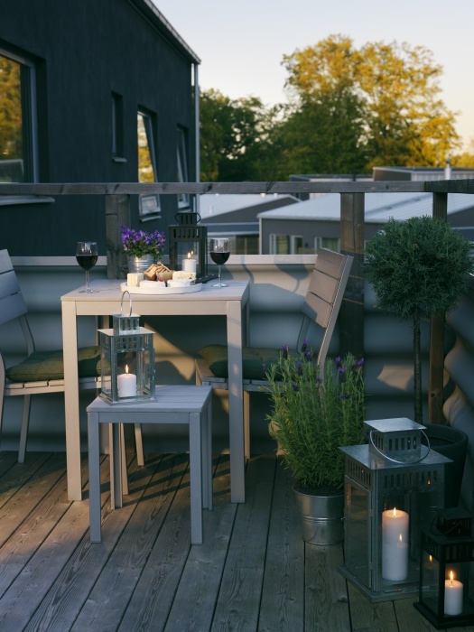 a small and cozy summer balcony with grey wooden furniture, potted greenery and blooms and candle lanterns