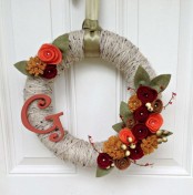 a bright and cool fall wreath covered with grey yarn, with fabric blooms, leaves and twigs and a monogram for a rustic feel
