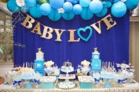a bold blue and gold dessert table with blue balloons and pompoms, a letter banner and bright blue and gold stands