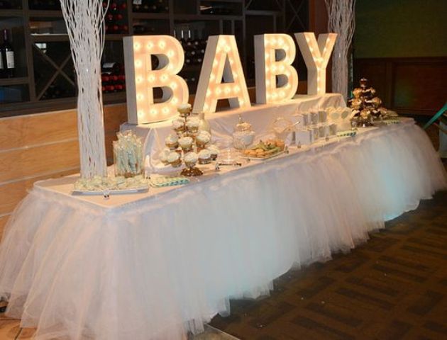 a bright white baby shower dessert table with lights, branch arrangements, a tulle tablecloth and lots of desserts