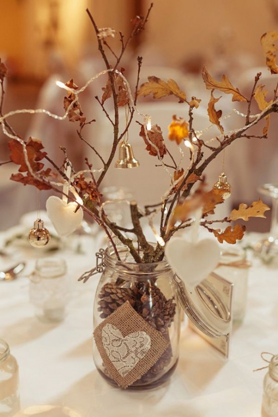 a rustic fall centerpiece of a glass vase with pinecones, branches with leaves and twigs, ornaments, bells and clay ornaments