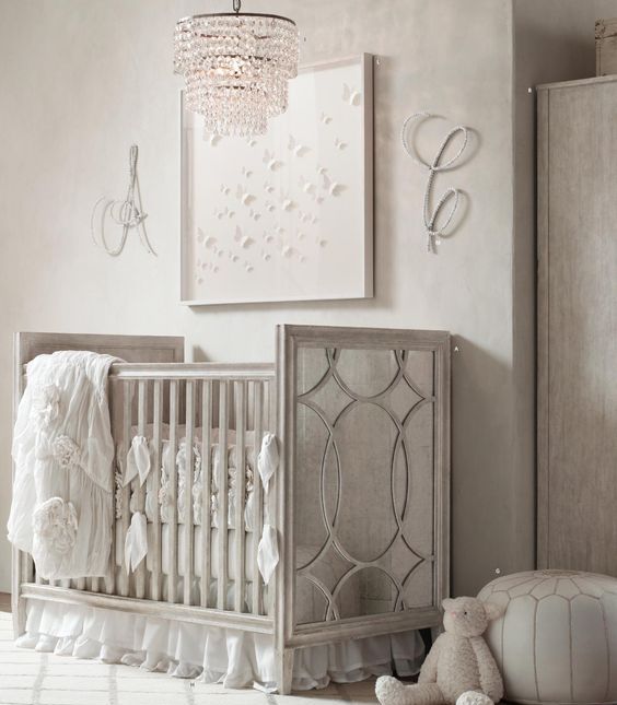 a vintage glam nursery in white and off whire, with a vintage crib, a pretty artwork and rhinestone monograms plus a crystal chandelier
