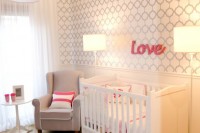 a simple modern nursery with a wallpaper accent wall, a white crib and lamps, a grye chair and a pink pendant lamp plus grey and white bedding