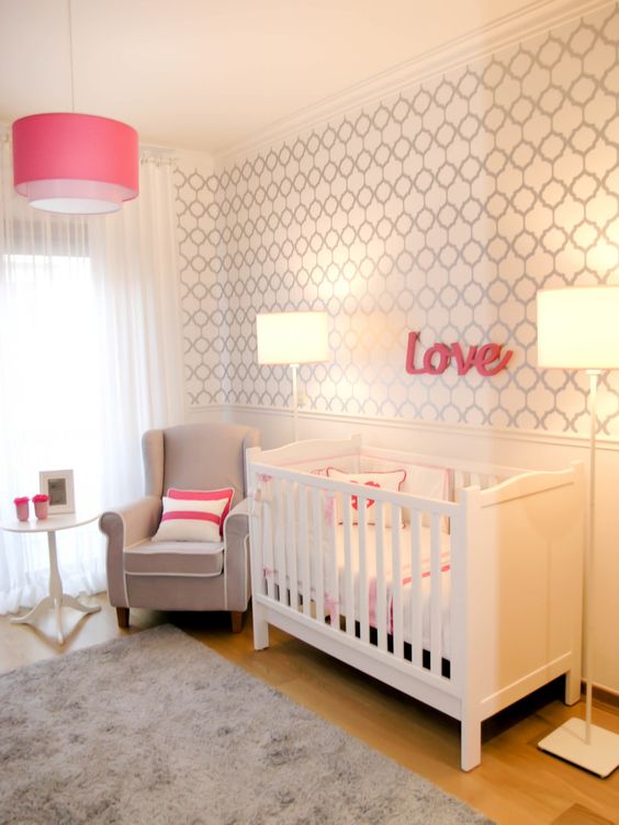 a simple modern nursery with a wallpaper accent wall, a white crib and lamps, a grye chair and a pink pendant lamp plus grey and white bedding