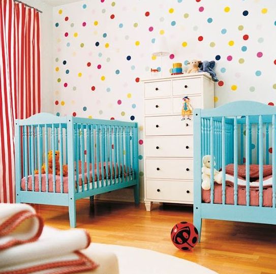 a colorful shared nursery with a bright polka dot wall, turquoise cribes, pink and white bedding and lots of toys