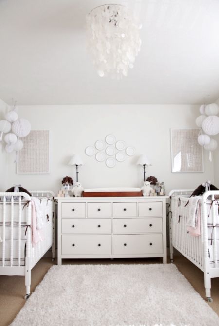 a gender neutral shared nursery with white furniture, white paper lamps, a mother of pearl chandelier and striped bedding