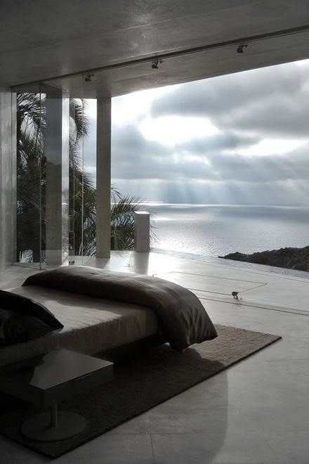 a minimalist bedroom done in grey shades and with a glass wall that opens up to a terrace with a lovely ocean view is a very dreamy space to be