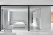 a super clean and minimalist bedroom in white, with an exposed brick wall, storage units and curtains is very airy and a glass wall connects it to the rest of the house
