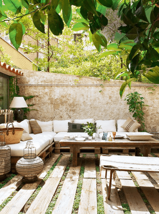 a relaxed outdoor Mediterranean space with a grass and wood plank floor, a pallet corner sofa with neutral upholstery, a wooden table and a daybed, some Moroccan lanterns