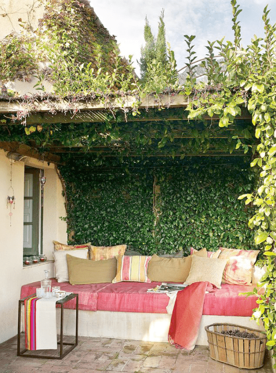 a welcoming Mediterranean space with greenery over it, with a built-in sofa with pink upholstery and pillows, a metal table with colorful towels and a basket with pinecones