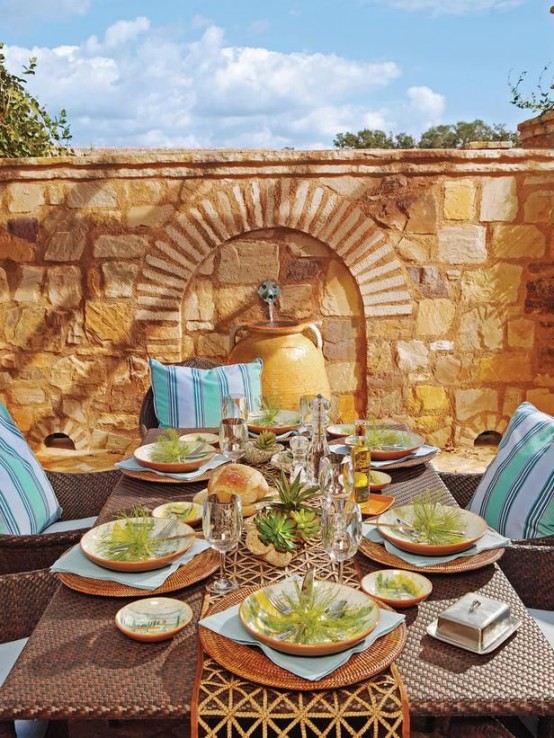 a gorgeous Mediterranean outdoor dining space with a stone wall with a fountain, a dark wicker dining seat, blue pillows, greenery and succulents for a fresh touch