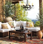 a Mediterranean otudoor space with a white corner sofa, a dark-stained coffee table, a vintage candle chandelier, greenery and vases around plus printed pillows