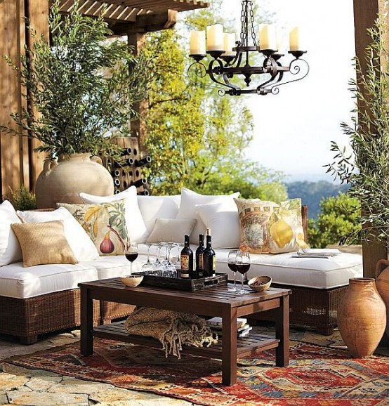 a Mediterranean otudoor space with a white corner sofa, a dark-stained coffee table, a vintage candle chandelier, greenery and vases around plus printed pillows