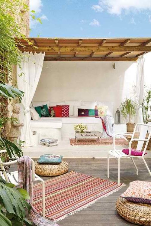 a beautiful and welcoming Mediterranean outdoor space with a white built in sofa with bright pillows, a roof over this seat, colorful rugs and pillows and white chairs and jute poufs