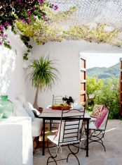a small outdoor dining area wiht a Mediterranean feel, with a white sofa, a wood and metal table, metal chairs with white cushions, bright blooms and pillows to add color