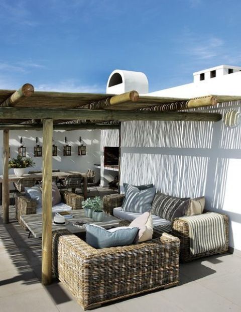 a neutral outdoor Mediterranean space with white walls, neutral wicker furniture with white upholstery, pastel pillows, potted greenery and blooms, lanterns on the wall